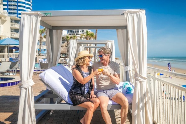 A couple relaxes on a beach cabana, enjoying drinks and a sunny day by the ocean.