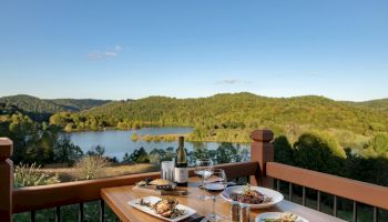 A wooden table set with dishes, a bottle of wine, and glasses overlooks a scenic view of a lake and green hills on a clear, sunny day.