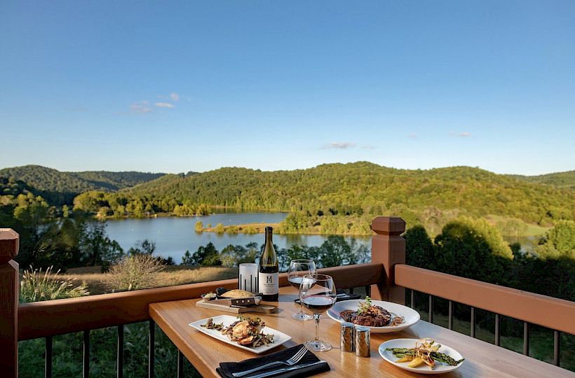 A wooden table set with dishes, a bottle of wine, and glasses overlooks a scenic view of a lake and green hills on a clear, sunny day.