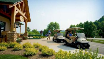 The image shows two people in golf carts with another person walking carrying golf clubs in front of a building surrounded by greenery and flowers.