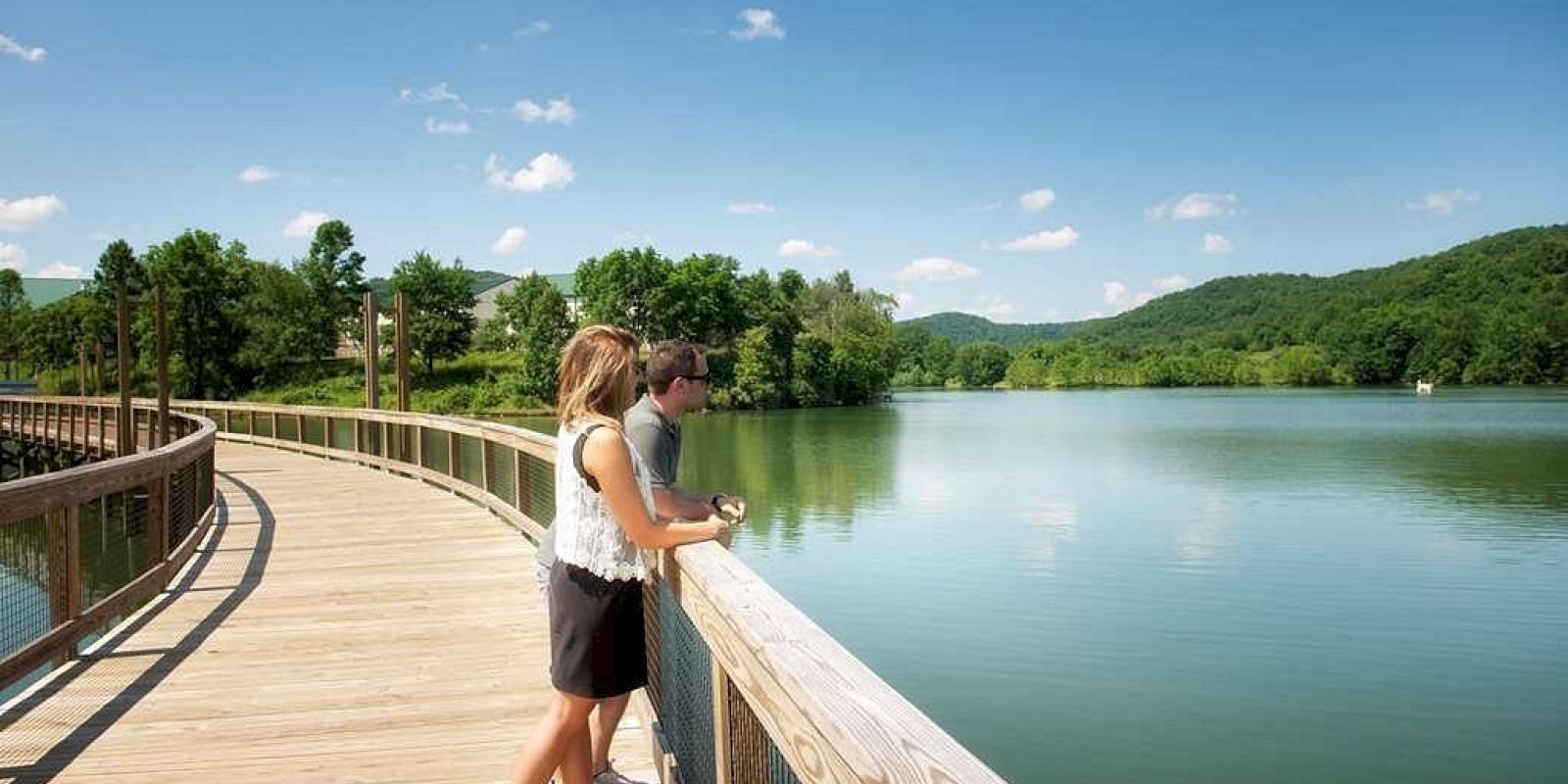 Two people stand on a wooden boardwalk overlooking a serene lake with green hills in the background, on a clear, sunny day.