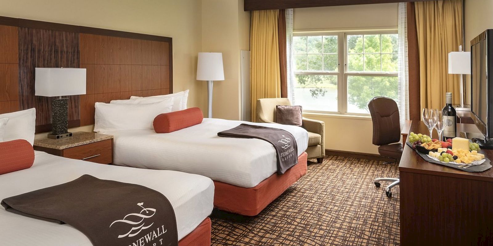 The image shows a hotel room with two double beds, a window with curtains, a desk with a TV, and a fruit and wine setup.