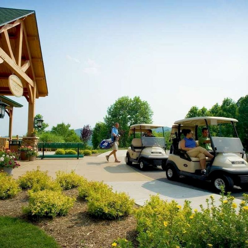 Two people are seated in golf carts near a building with wooden beams. Another person walks nearby with a golf bag. Flowers are in the foreground.