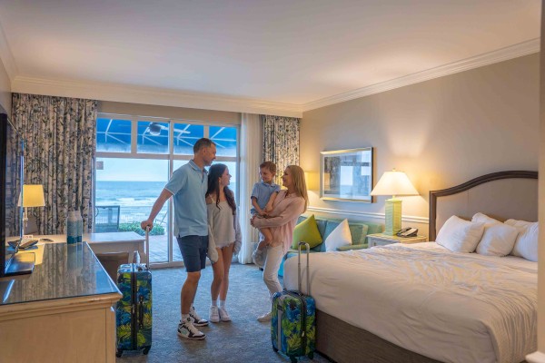 A family of four stands in a hotel room with luggage, looking out a window that shows an ocean view, suggesting a relaxed vacation setting.