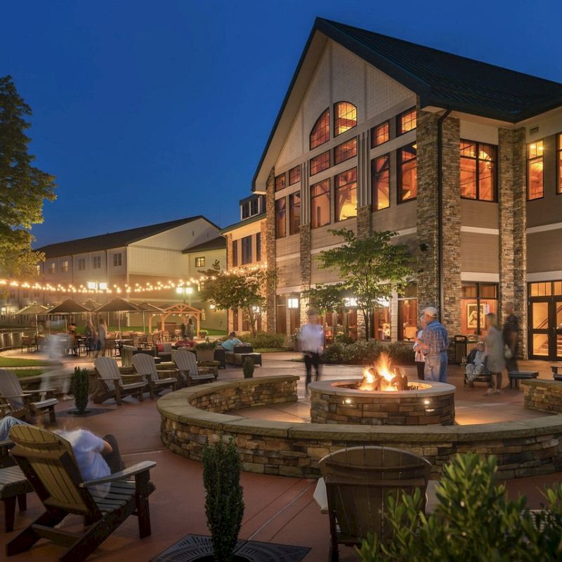 The image depicts a well-lit building with large windows and an outdoor seating area surrounding a fire pit. String lights add ambiance.