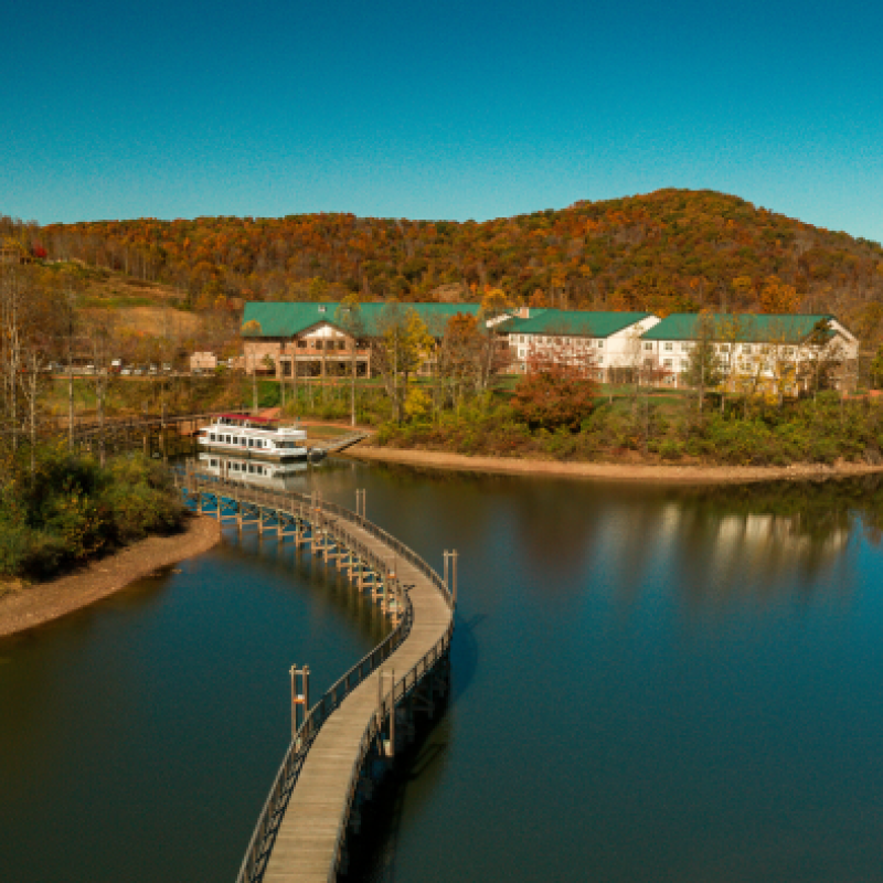 A scenic view of a lake with a curved boardwalk, leading to a building complex with green roofs, surrounded by autumn-colored trees and hills.