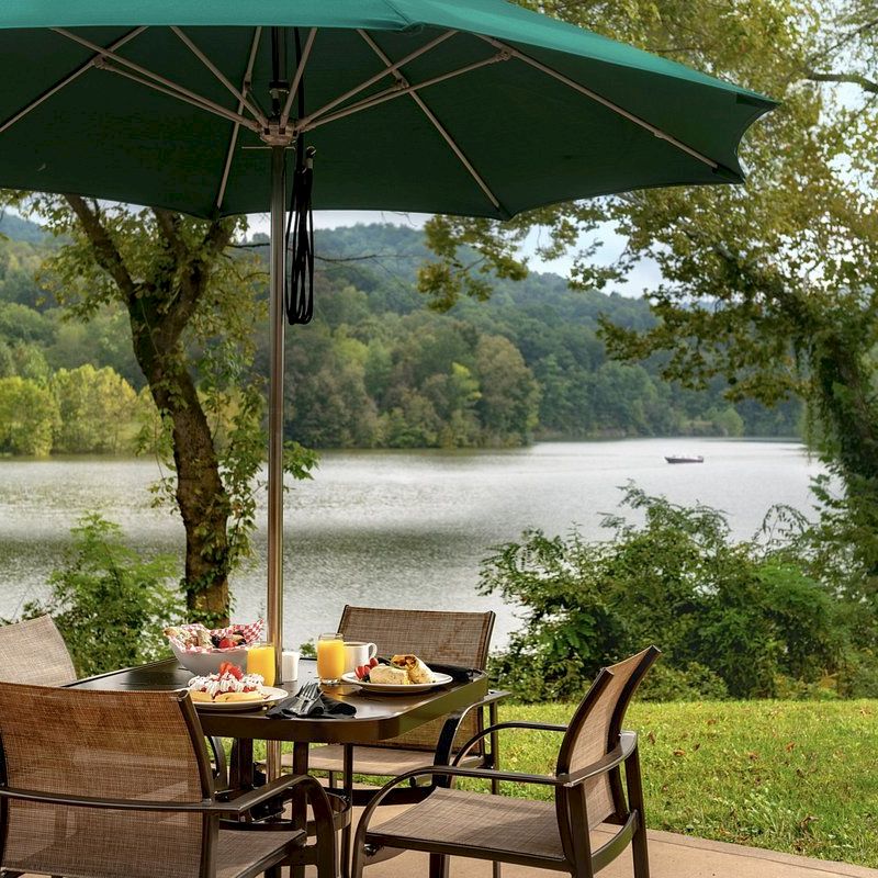 A patio set with food under a green umbrella, overlooking a lake and surrounded by lush trees; a relaxing dining scene.