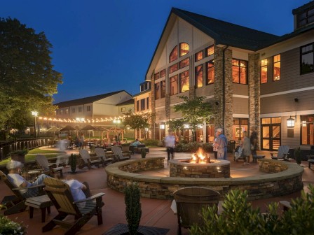 A nighttime scene of a large, well-lit building with an outdoor seating area featuring a fire pit, string lights, and people socializing.