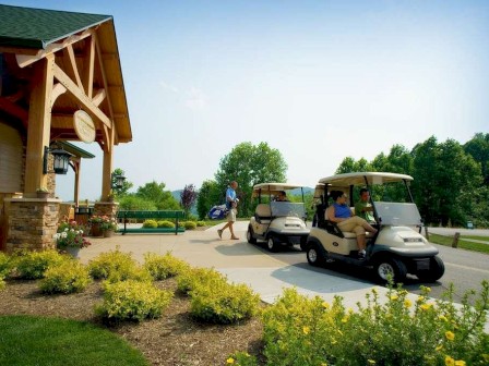 The image shows two golf carts parked near a wooden building with a person walking, carrying a golf bag, and a landscaped area with yellow flowers.