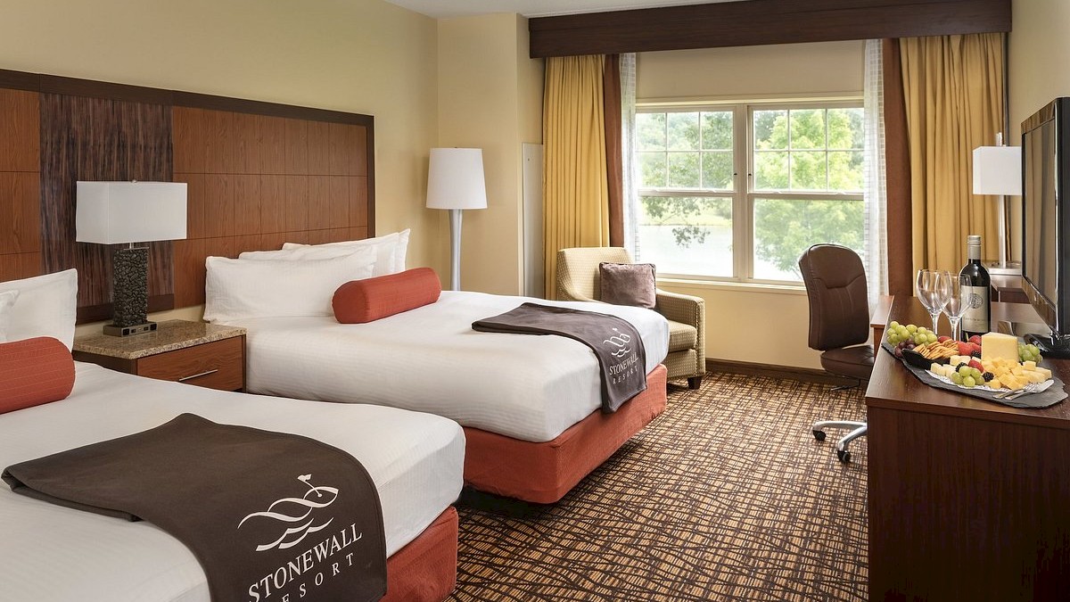 A hotel room with two double beds, a TV, a window with a view, armchair, floor lamp, and a table with wine and a fruit plate.