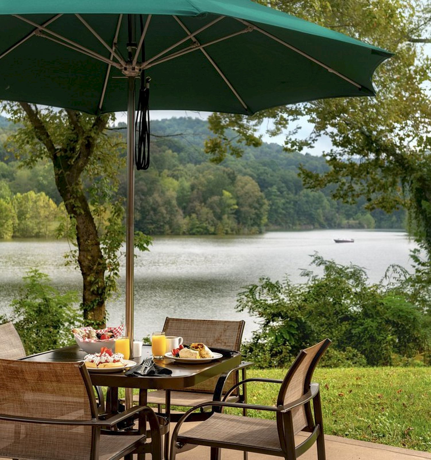 Outdoor dining setup with a green umbrella, table, and chairs, overlooking a serene lake with hills in the background. End the sentence.