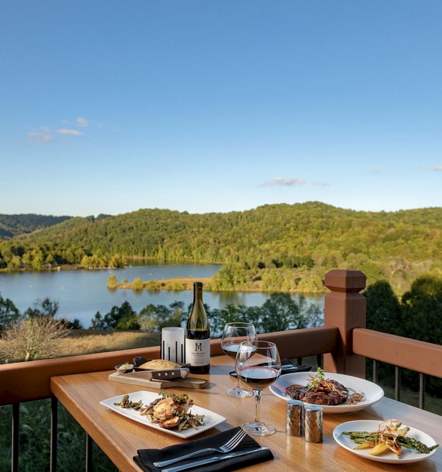 A dining table with plates of food, wine, and glasses overlooks a scenic landscape of a lake surrounded by green hills under a clear blue sky.