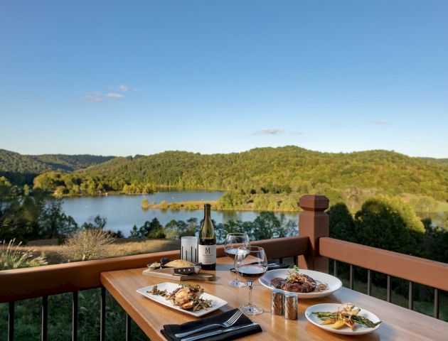 A dining table set with food and wine overlooks a scenic landscape with a lake and lush green hills under a clear blue sky.