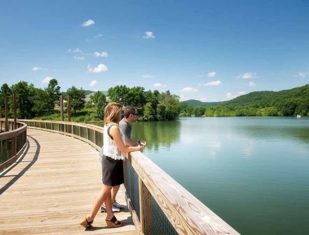 A couple stands on a wooden boardwalk overlooking a serene lake surrounded by lush green hills under a clear blue sky, enjoying the view.