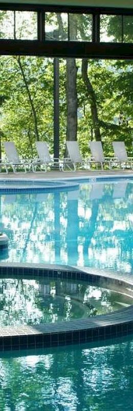 An indoor pool area with large glass windows, a hot tub, and an adjacent outdoor pool with lounge chairs in the background.