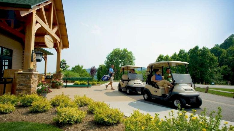 The image shows people at a golf clubhouse with two golf carts parked on a pathway, surrounded by greenery and small yellow flowers.