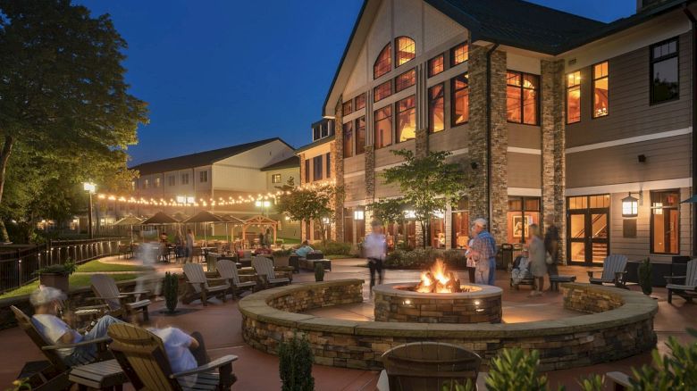 The image shows a cozy outdoor patio area of a building at twilight. People are gathered around a fire pit with string lights overhead, creating a warm ambiance.