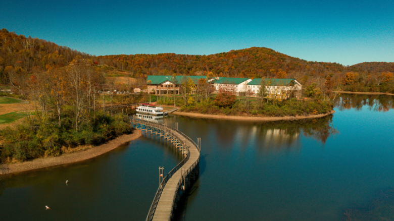 An aerial view shows a winding bridge leading to buildings surrounded by trees, with a boat docked nearby on a calm lake, under a clear sky.
