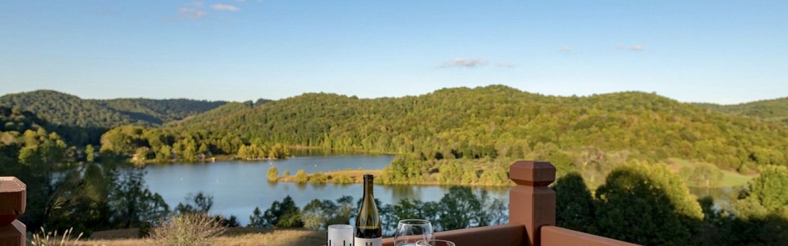 A wooden table set with plates of food, wine, and glasses overlooks a scenic lake surrounded by lush green hills under a clear blue sky.
