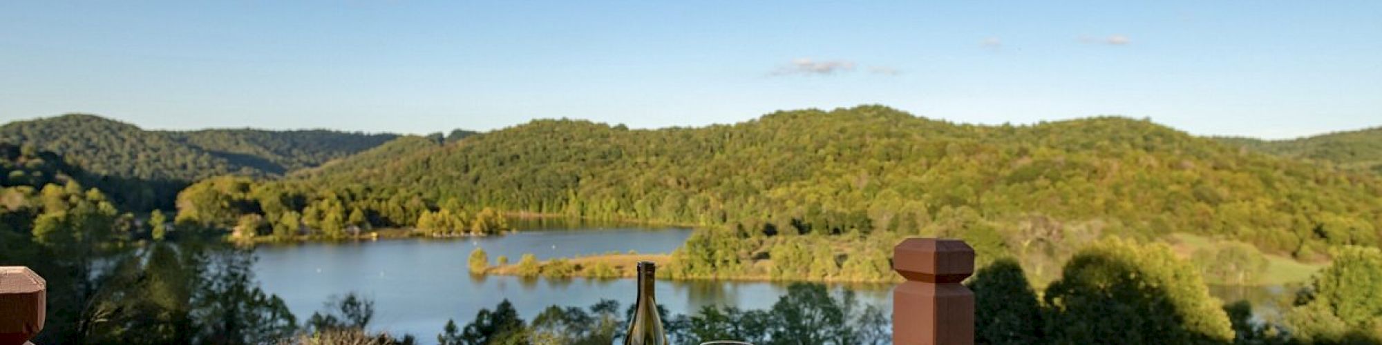 A wooden table set with plates of food, wine, and glasses overlooks a scenic lake surrounded by lush green hills under a clear blue sky.