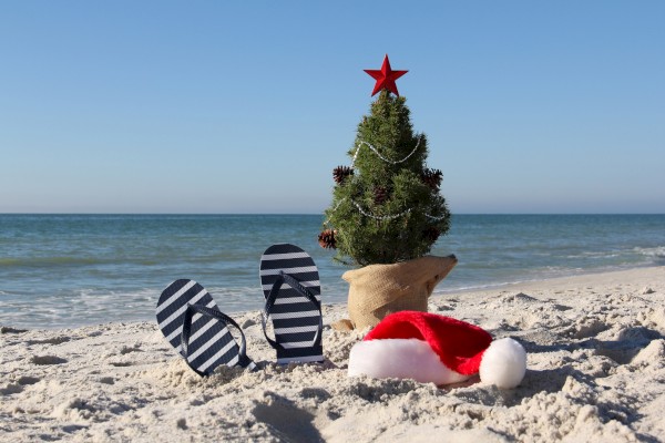 A small Christmas tree, striped flip-flops, and a Santa hat are on a sandy beach with the ocean in the background.