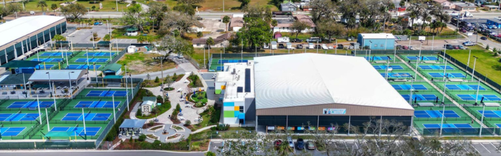 An aerial view of a driving range and practice facility with multiple netted bays, a central clubhouse, and surrounding parking.