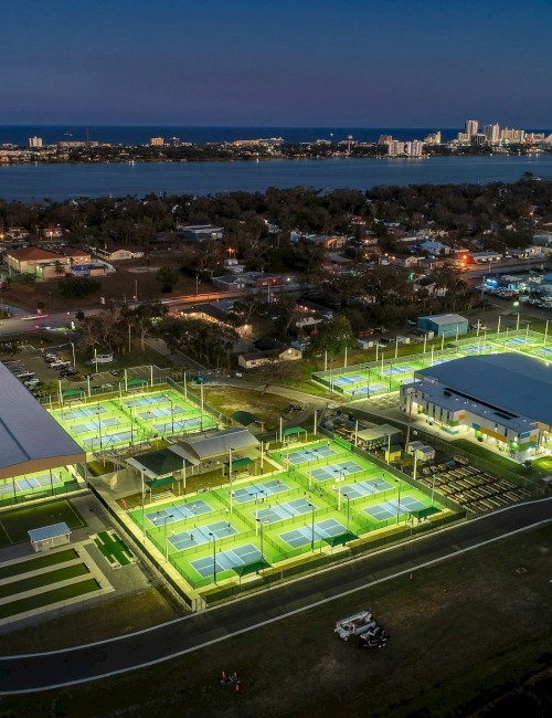 Aerial view of a well-lit sports complex at dusk, with multiple illuminated tennis courts and a large building, near a coastal city shoreline.