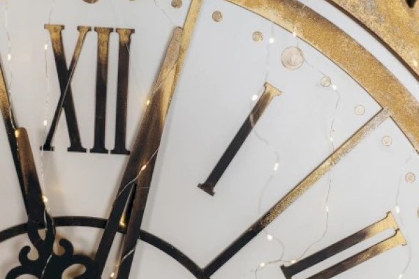 A close-up of a vintage clock with white dial, gold numerals, and black hands, showing Roman numerals XII, I, VII, and decorative details.