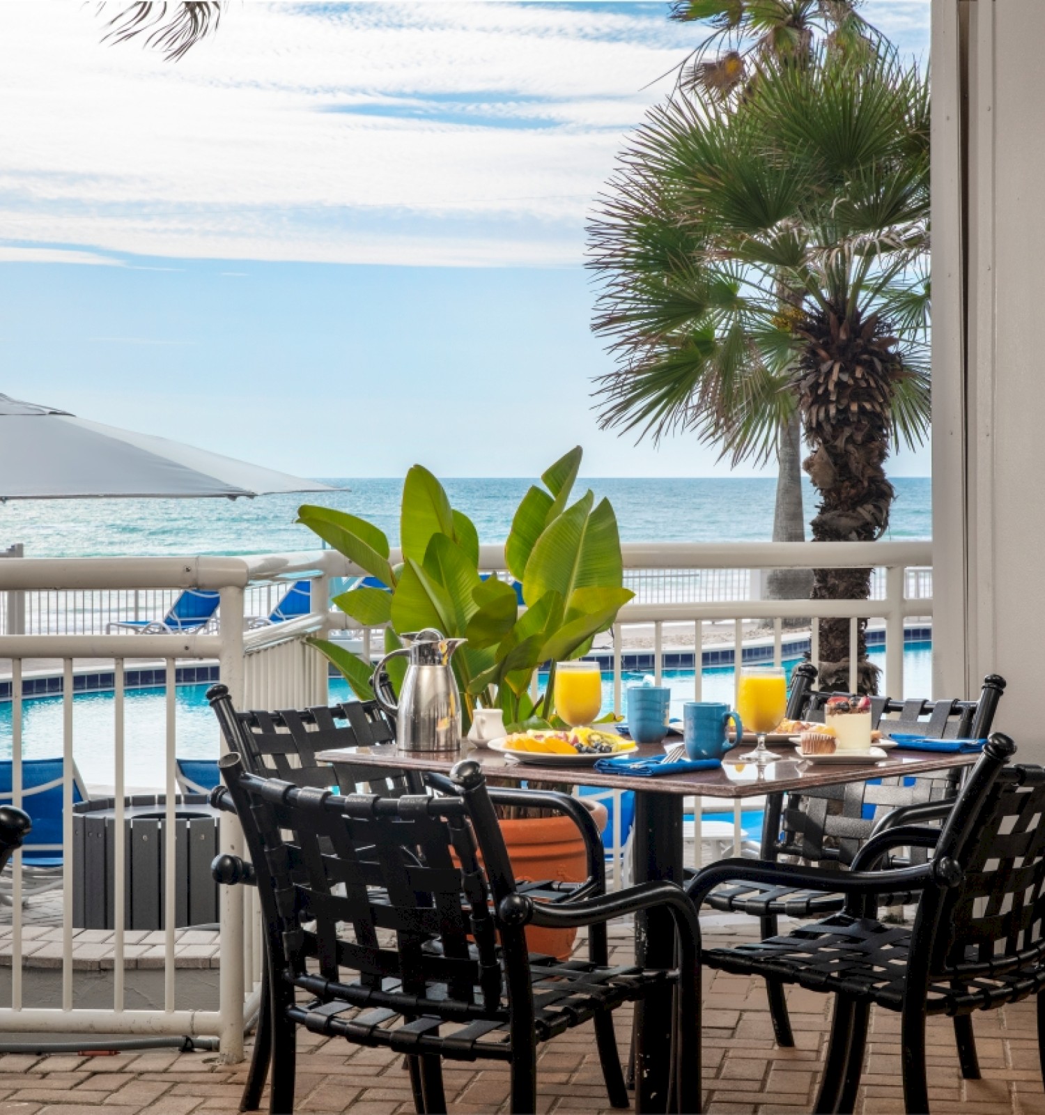 A sunny seaside dining setup on a balcony with black wicker chairs, a few tables with plates and drinks, palm trees, and a view of the ocean and umbrellas.