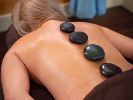 A person lying face down getting a hot stone spa massage with smooth stones along the back, dim cozy room, and towels.