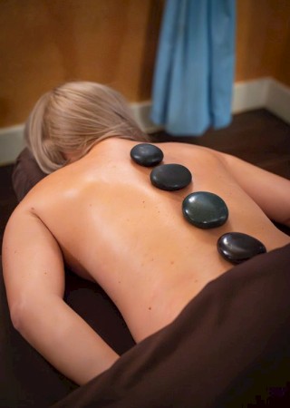 A person lying face down getting a hot stone spa massage with smooth stones along the back, dim cozy room, and towels.