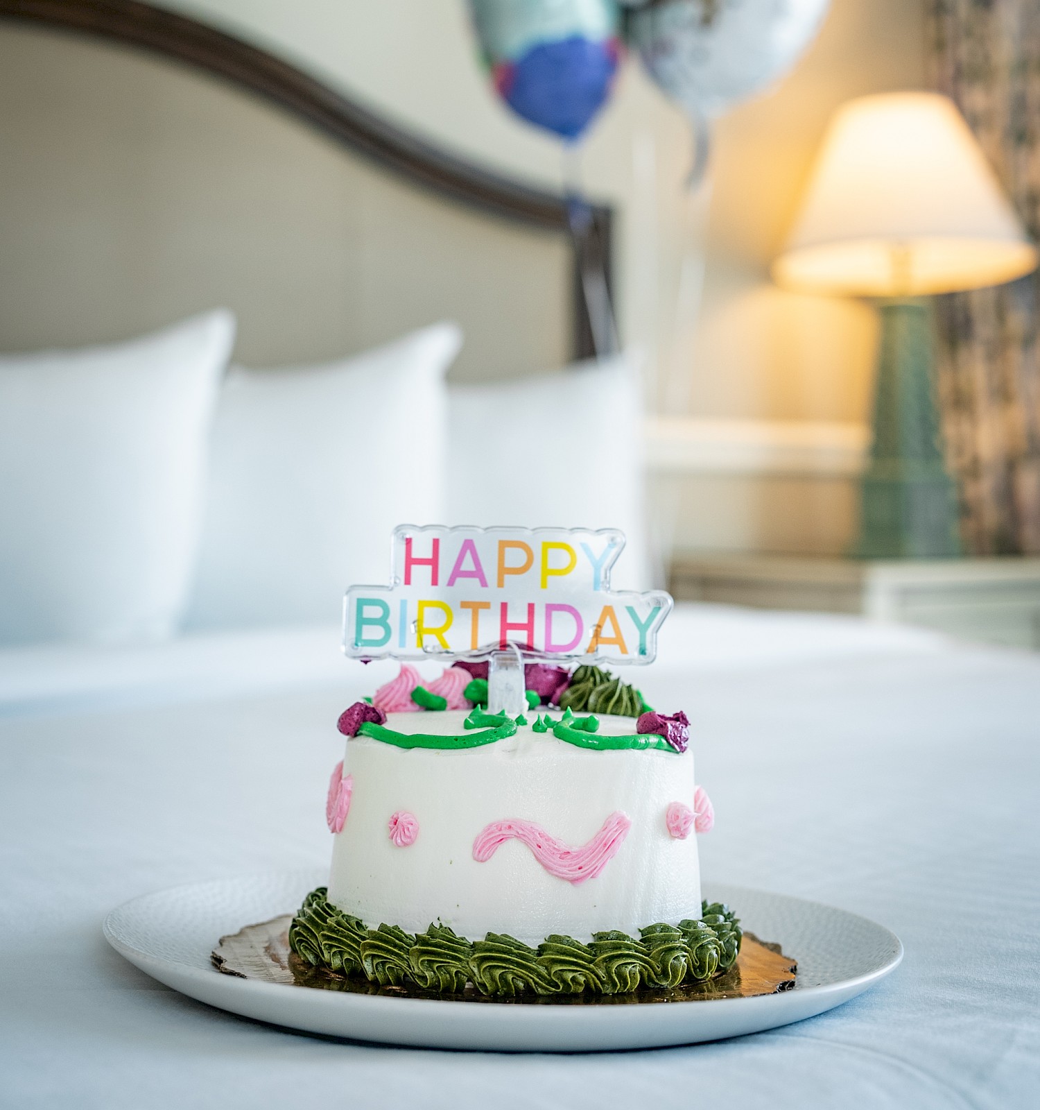 A birthday cake with pink icing, colorful “Happy Birthday” topper, green leaves border, and balloons in a cozy bedroom backdrop.