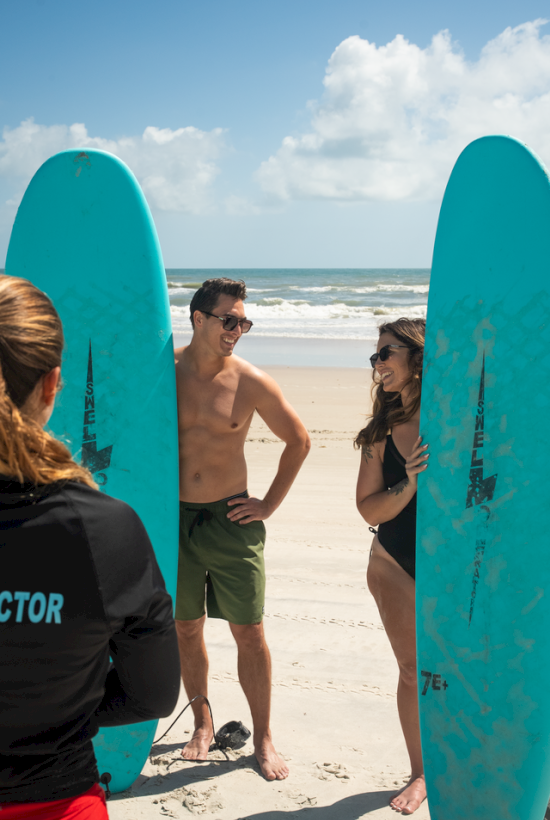 Two smiling people holding surfboards and chatting with a surf instructor on a sunny beach.