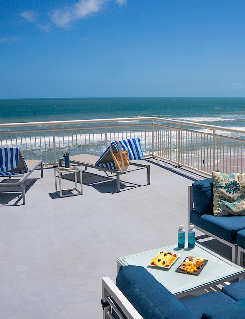 A balcony with blue furniture, towels, drinks, and snacks overlooking a beach and ocean under a blue sky.