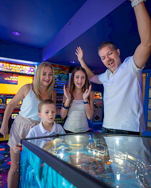A family plays arcade games at a bowling alley or arcade room; kids and adults smile, with a high-energy, celebratory vibe.