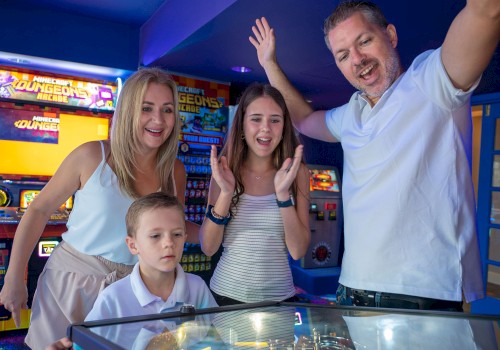 A family plays arcade games at a bowling alley or arcade room; kids and adults smile, with a high-energy, celebratory vibe.