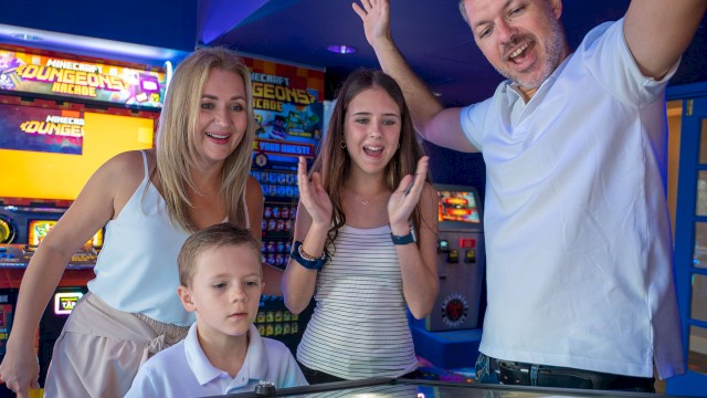 A family plays arcade games at a bowling alley or arcade room; kids and adults smile, with a high-energy, celebratory vibe.